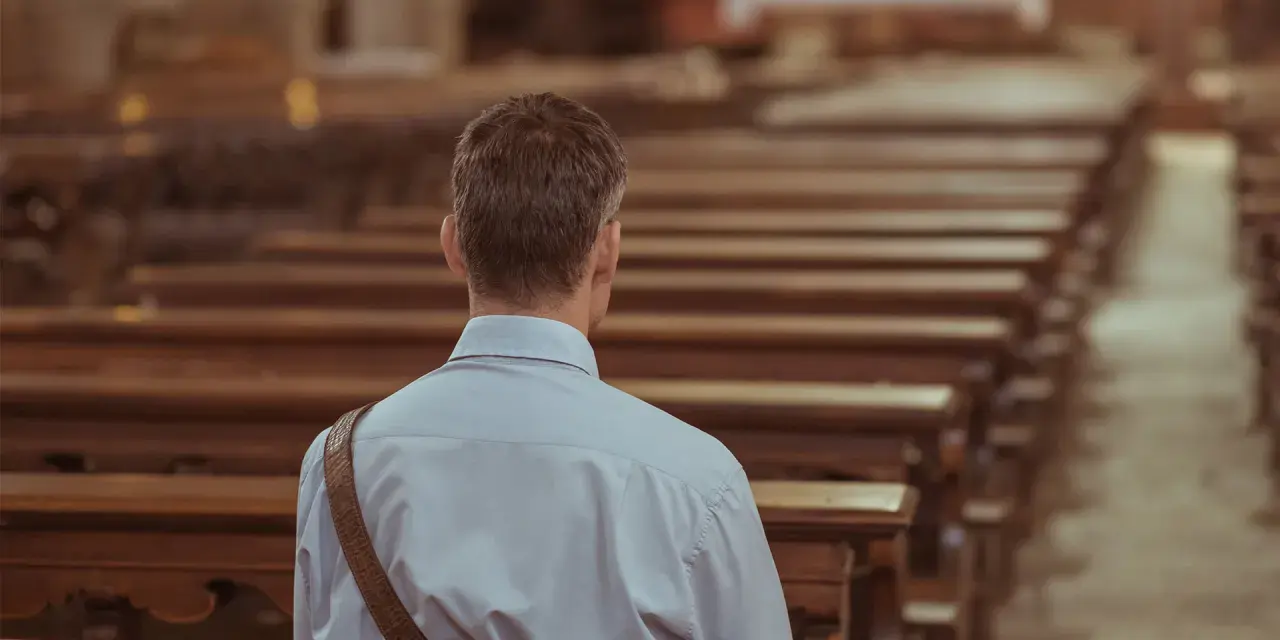 Mężczyzna siedzi w pustej ławce kościelnej, pogrążony w zadumie. To chwila wyciszenia i duchowości.