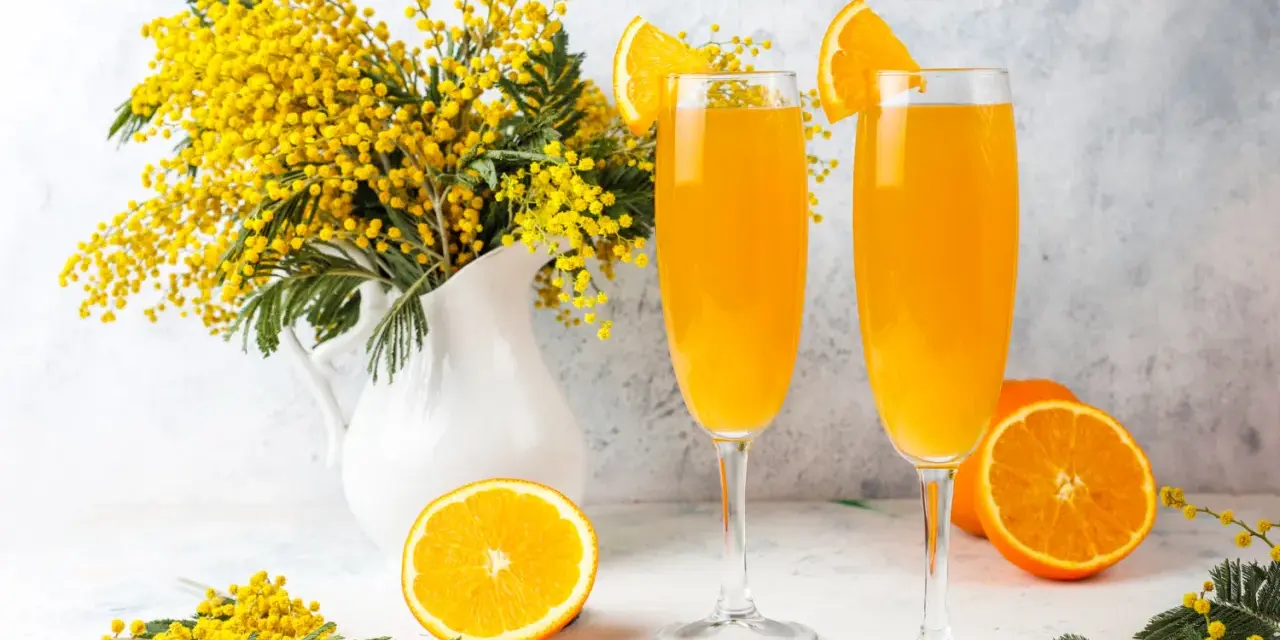 Dwa kieliszki z pomarańczowym napojem, ozdobione plasterkami pomarańczy. Obok bukiet mimozy i połówka pomarańczy. Idealne na wiosenny mimoza drink przepis.