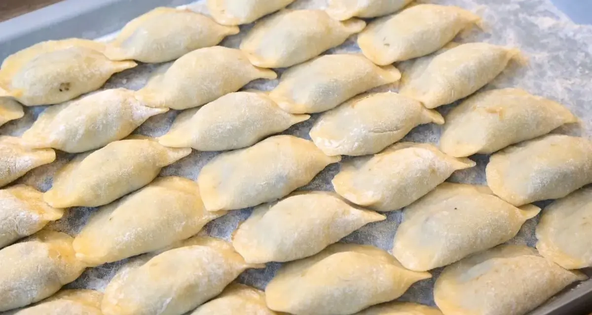 Ułożone na tacy surowe pierogi, posypane mąką.