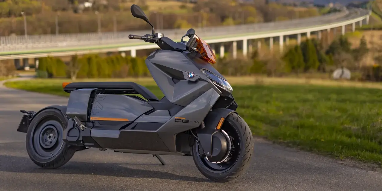 Szary skuter elektryczny BMW CE 04 na tle malowniczego krajobrazu z mostem i wzgórzami.