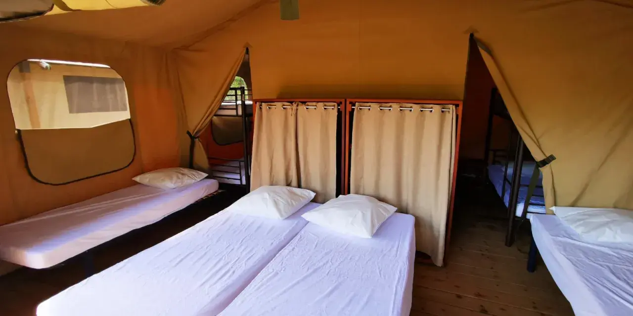 Intérieur d'une chambre de colonie de vacances avec plusieurs lits simples, draps blancs et oreillers. Des rideaux séparent les espaces.