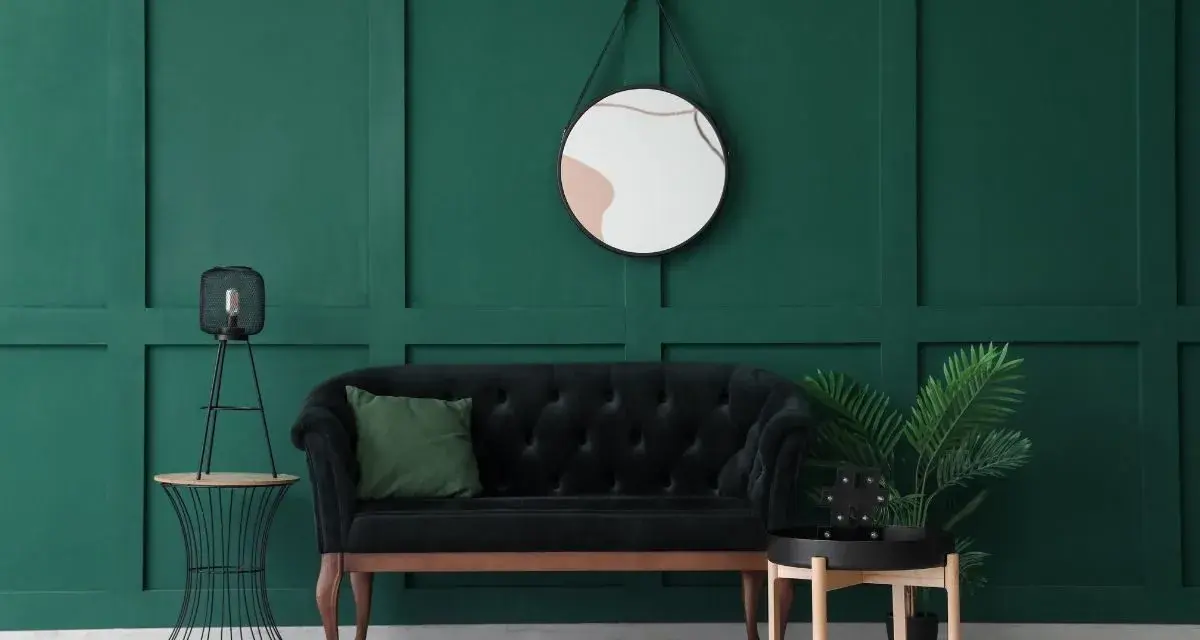 Un elegante divano nero con cuscino verde, un tavolino con lampada e una pianta, e uno specchio dietro il divano su una parete verde scuro.