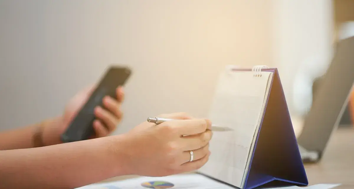 Ręce planują zadania na kalendarzu, trzymając telefon. Zapewnia to ciągłość pracy i organizację dnia.
