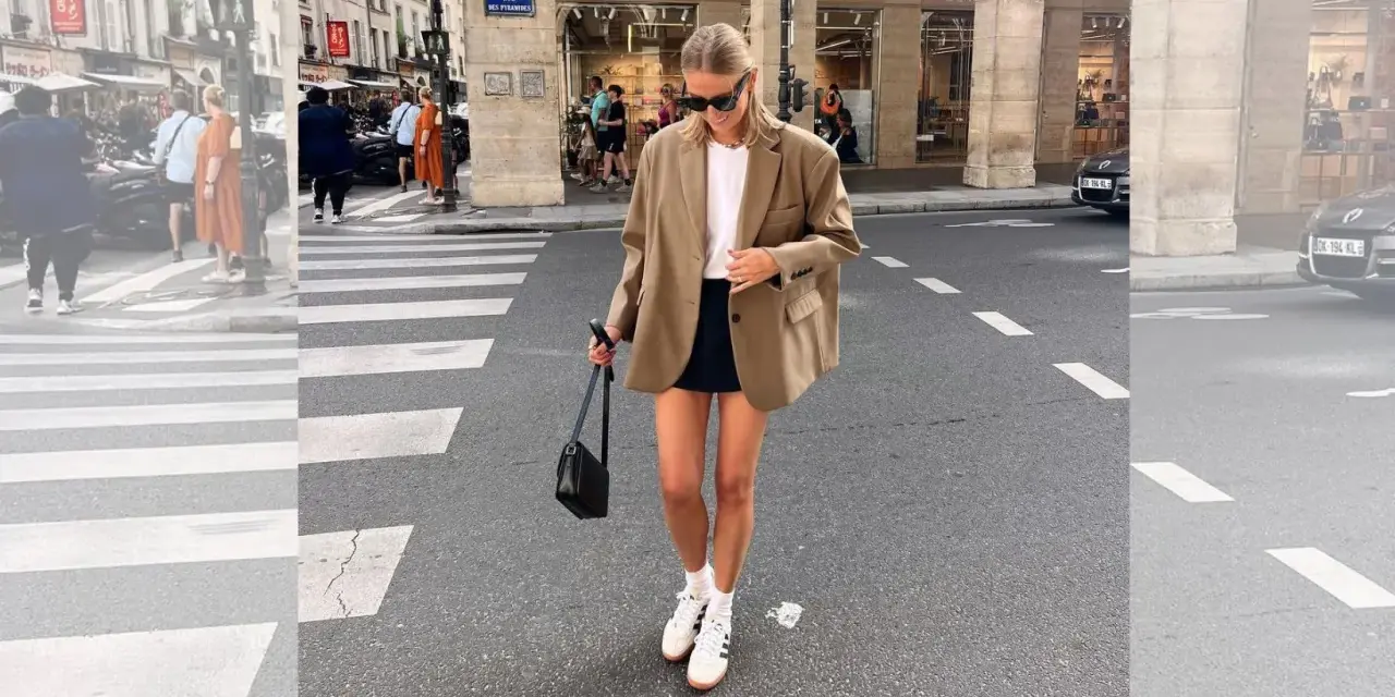 Mujer con look con blazer beige, falda negra y zapatillas blancas cruza un paso de peatones en París.