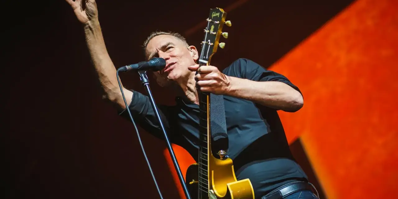 Bryan Adams na koncercie, w czarnej koszuli, z gitarą i mikrofonem, wznosi rękę w geście pozdrowienia.