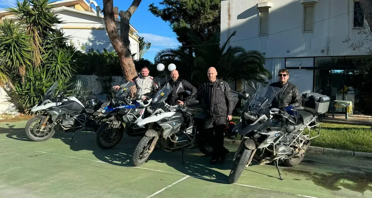 Czterech motocyklistów w pełnym rynsztunku gotowych na **prom na Sycylię**. Motocykle BMW GS zaparkowane na zielonym placu, w tle budynek i palmy.