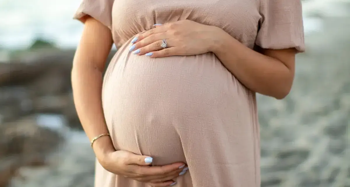 Brzuszek ciężarnej kobiety w beżowej sukience, dłonie delikatnie go obejmują. Warto pamiętać o bezpieczeństwie i unikać stosowania **retinolu w ciąży**.