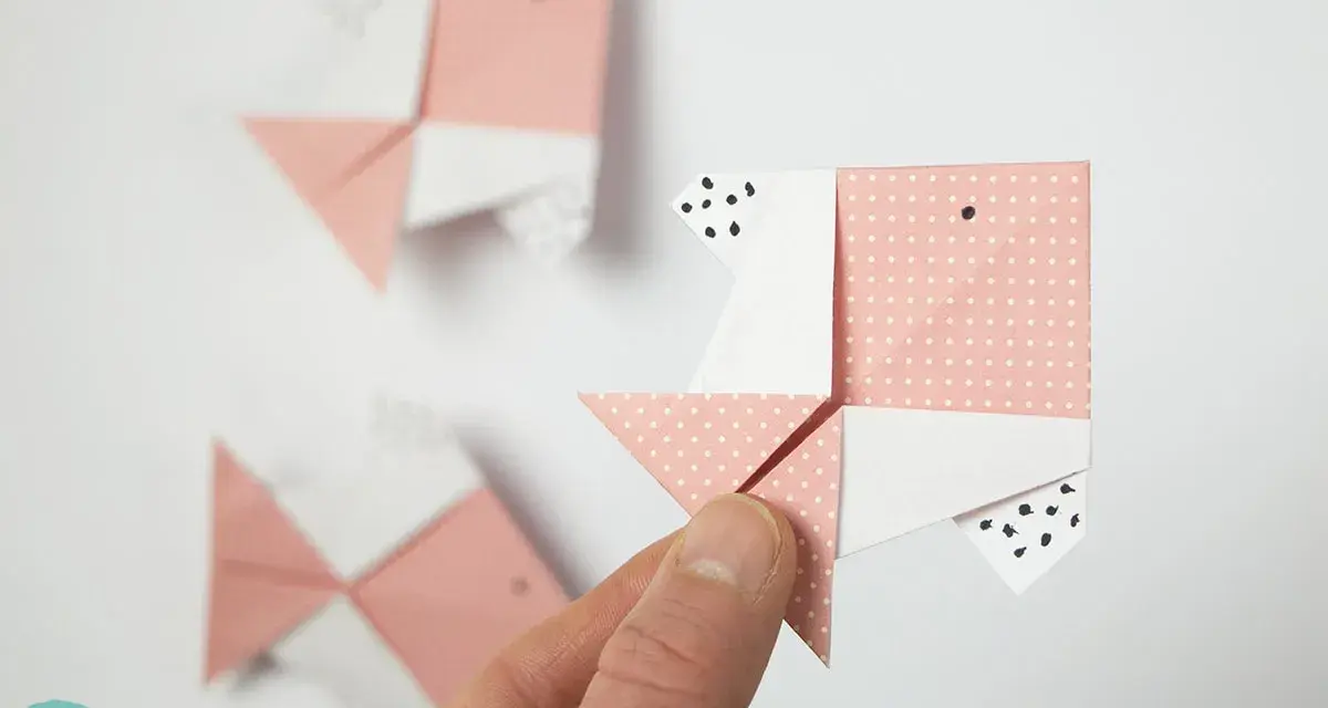 Hand hält Origami-Fisch. Mehrere rosa und weiße Papierfische, die gefaltet wurden, sind im Hintergrund zu sehen.