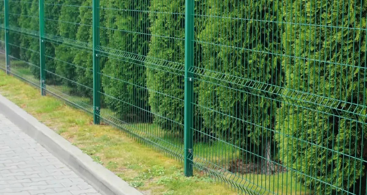 Zielone, metalowe ogrodzenie panelowe, jeden z popularnych rodzajów ogrodzeń, oddziela chodnik od gęstego żywopłotu.
