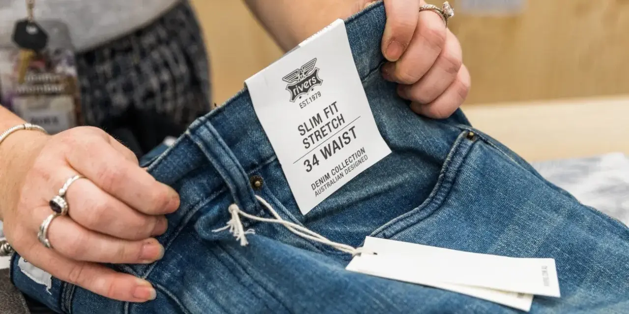Dłonie trzymają niebieskie jeansy z metką "SLIM FIT STRETCH 34 WAIST". Idealny rozmiar jeansów dla Ciebie.