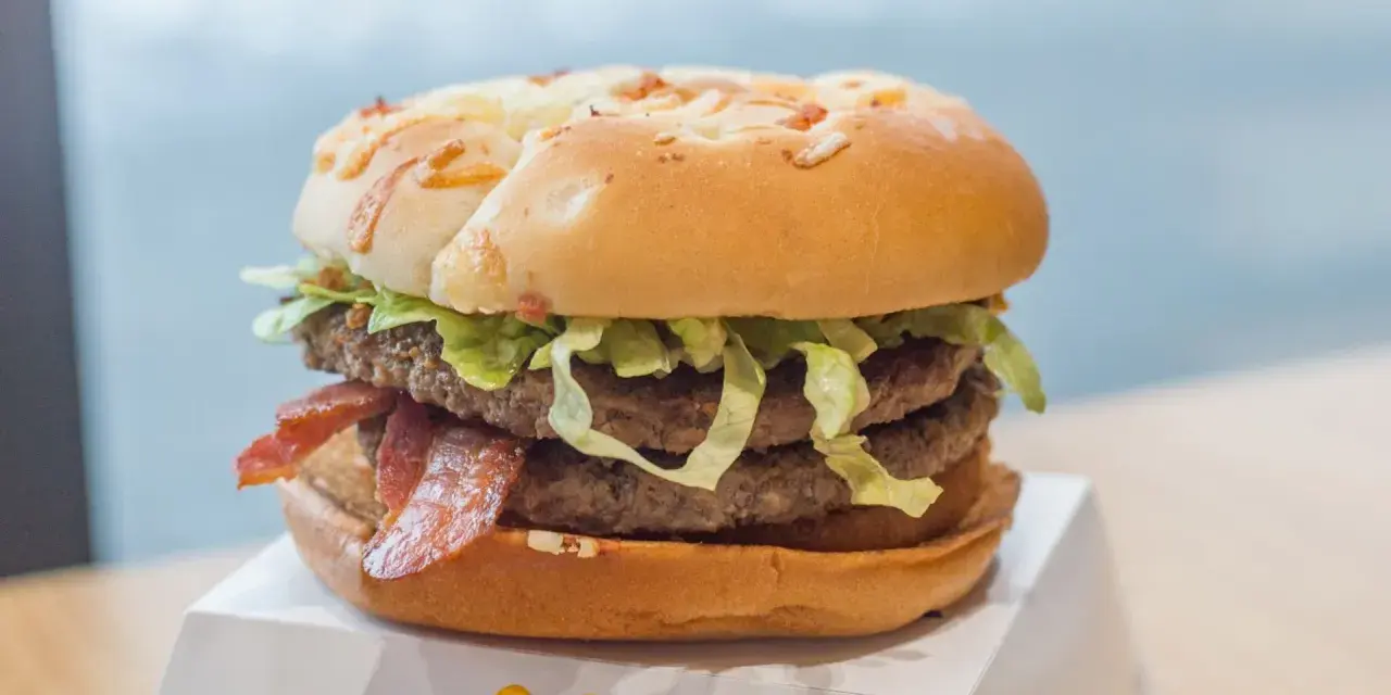 Duży burger z dwoma kotletami, bekonem i sałatą na białym opakowaniu.