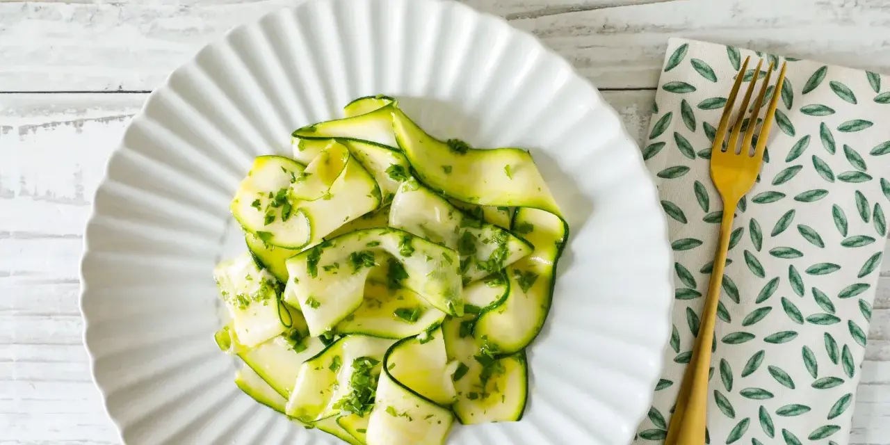Delicato carpaccio di zucchine condito con erbe fresche, servito su un piatto bianco con forchetta dorata.