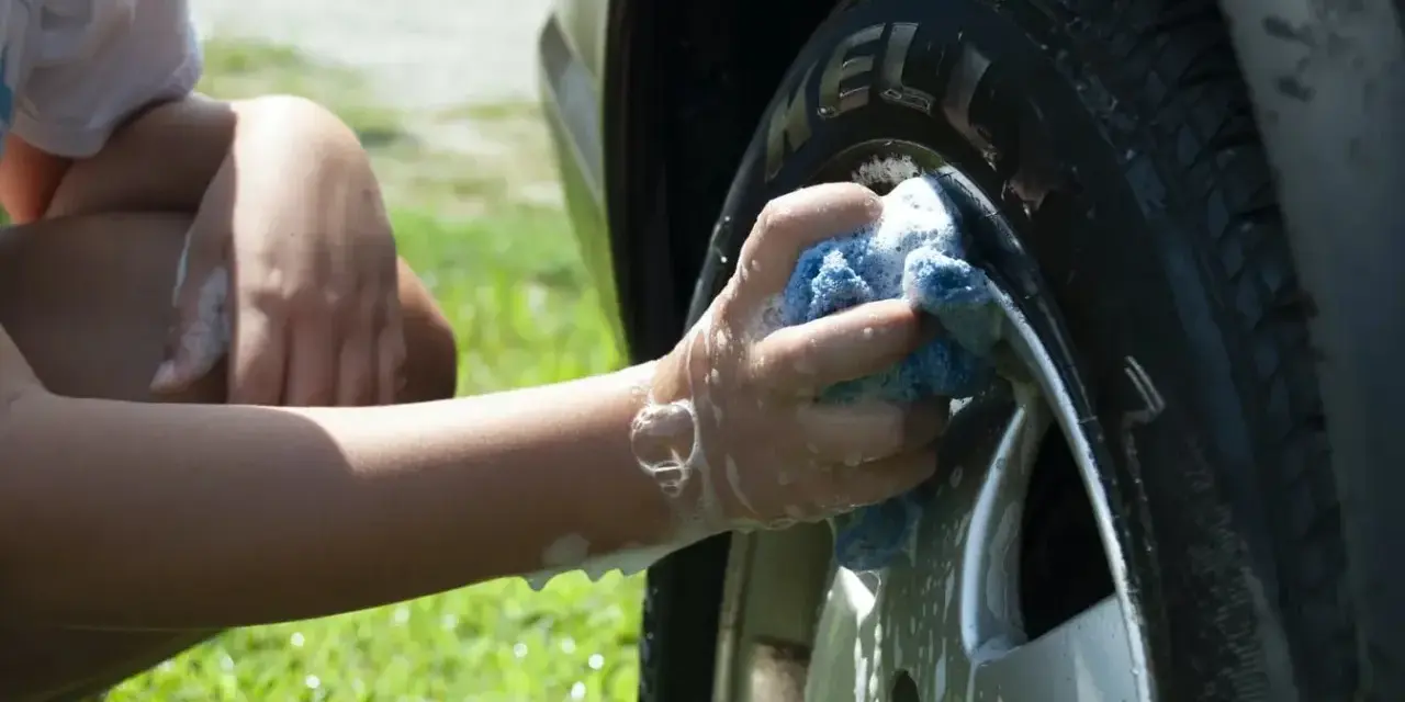 A person's arm and hand, covered in suds, scrub a car tire with a blue sponge. It seems they are testing if you can use dish soap to wash your car.