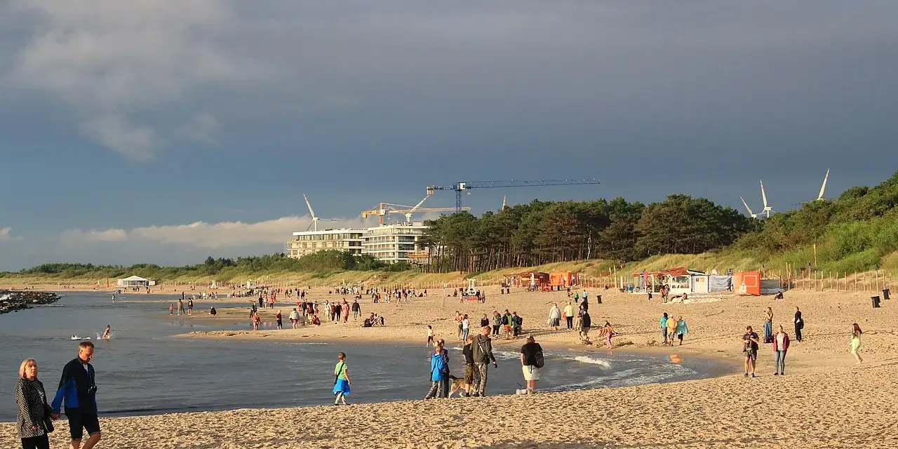 Nadmorskie miasto nad Wieprzą tętni życiem. Ludzie spacerują po plaży, bawią się w wodzie, a w tle widać budowę i wiatraki.