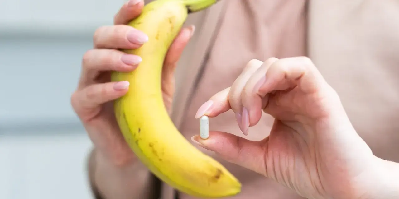 Kobieta trzyma banana i tabletkę, zastanawiając się, ile potasu dziennie powinna przyjmować.