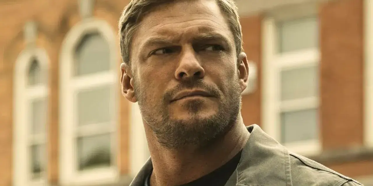 Alan Ritchson jako Jack Reacher. W tle ceglany budynek z białymi oknami.