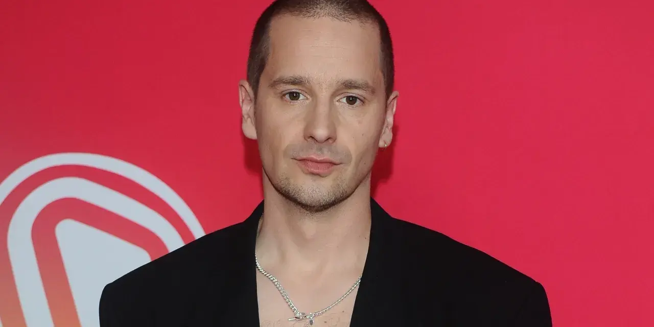 Krzysztof Zalewski w czarnym garniturze, z naszyjnikiem, na tle czerwonym z białym logo.