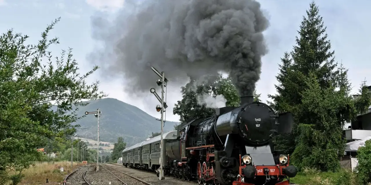 Czarny, parowy pociąg retro z wagonami, wydobywający kłęby dymu, jedzie po torach wśród zieleni i gór.