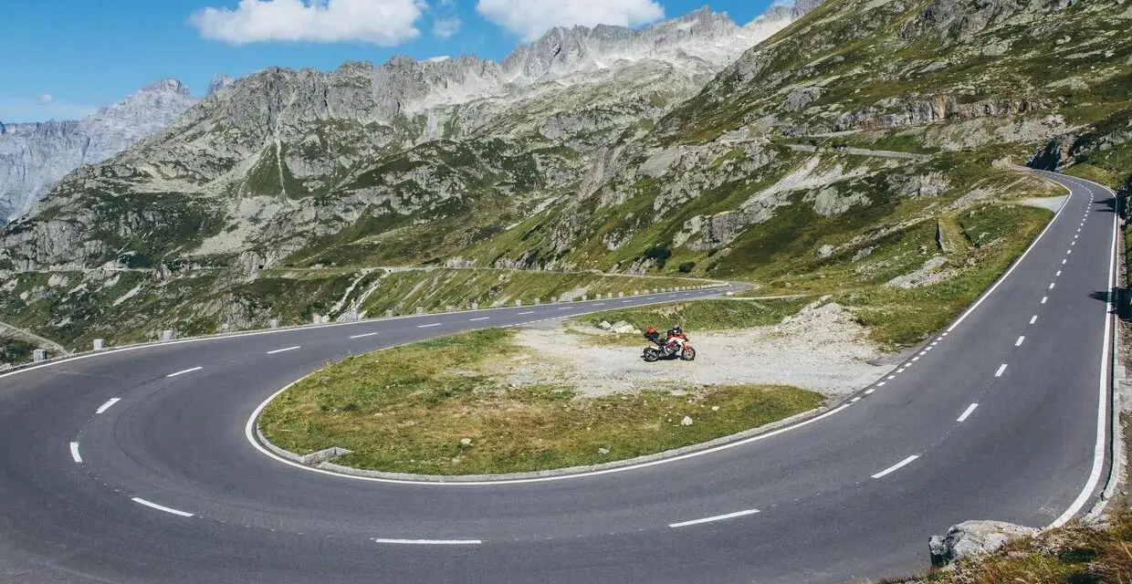 Una moto attende su un tornante del Grimsel Pass, tra montagne verdi e cielo azzurro.