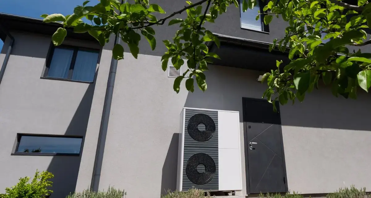 Unità esterna di una pompa di calore, moderna e discreta, installata accanto a una porta su una casa grigia.
