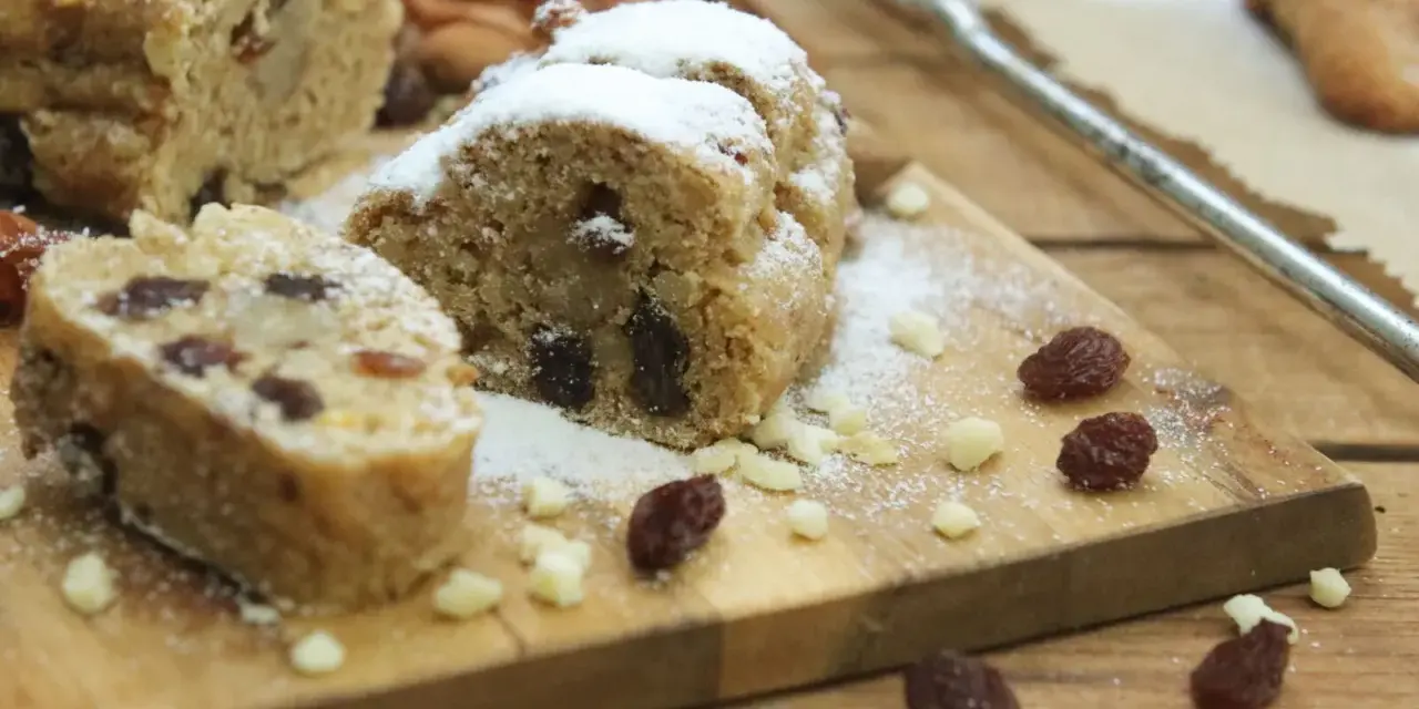 Stollen formen ohne backform: saftige Stollenstücke mit Rosinen und Puderzucker auf Holzbrett, umgeben von Nüssen und Trockenfrüchten.