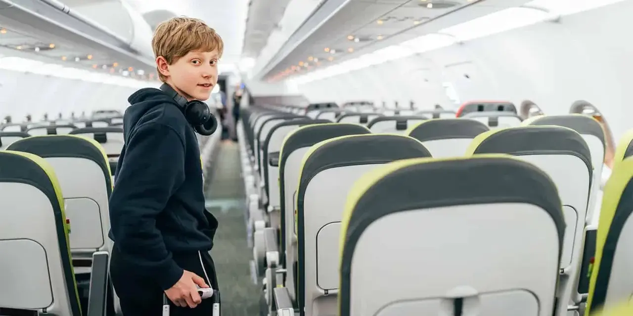 Chłopiec z słuchawkami na szyi i walizką w ręku stoi w przejściu samolotu. Od ilu lat można latać samemu? Ryanair pozwala na samodzielne podróże od 12 roku życia.