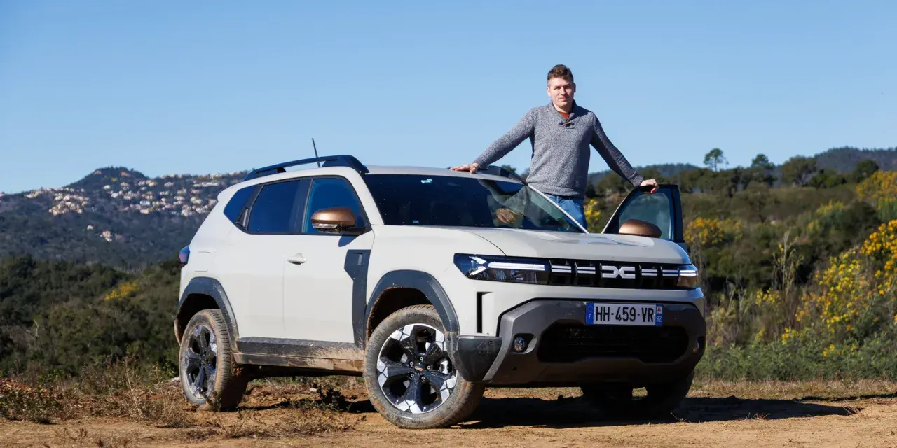 Mężczyzna opiera się o maskę jasnoszarego Dacia Duster, hybrydy z fabrycznym LPG, na tle wzgórz.
