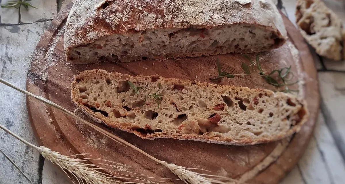 Chleb z suszonymi pomidorami, pokrojony na desce, z gałązką rozmarynu i kłosami zbóż.