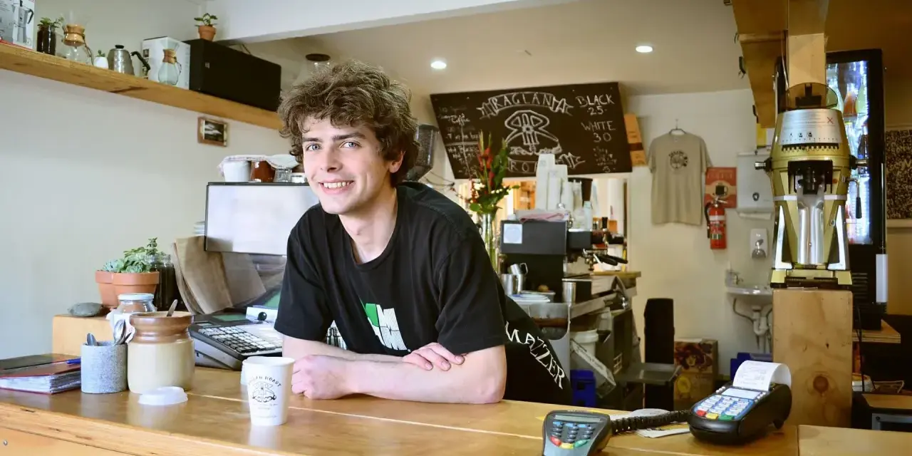 Ein junger Mann mit lockigen Haaren lächelt hinter der Theke eines Cafés. Er scheint seine **zwei Werkstudentenjobs** zu genießen, während er Kaffee zubereitet.