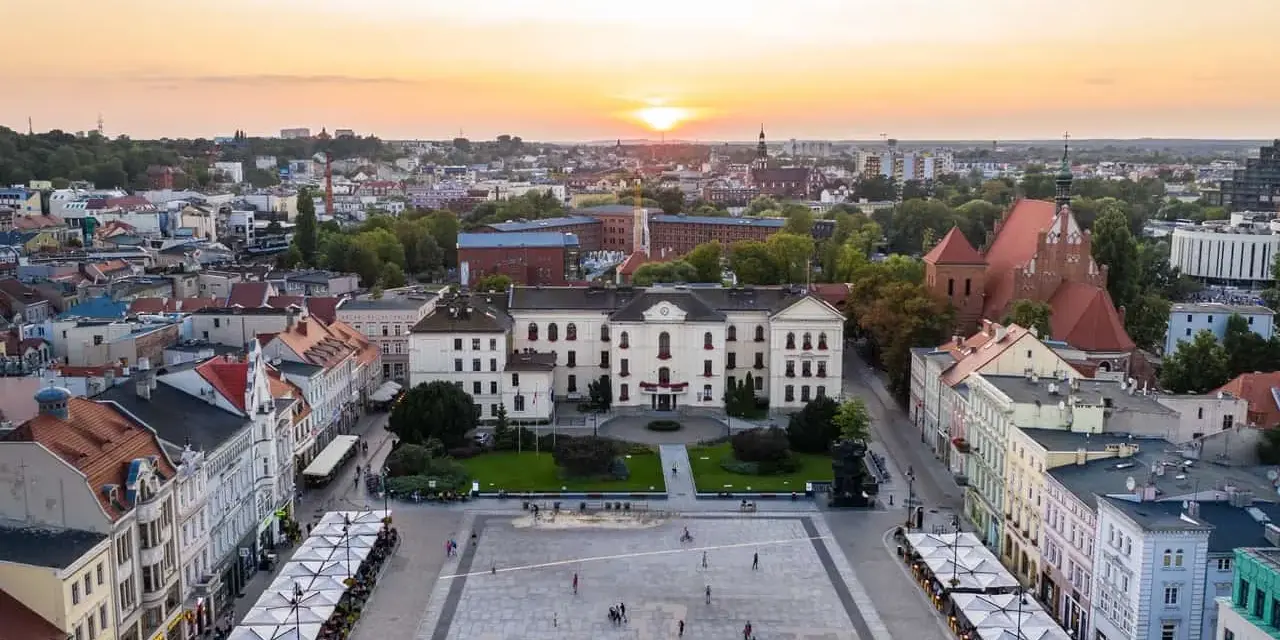 Zachód słońca nad rynkiem, gdzie widać piękną pierzeję kamienic i ratusz. Ludzie spacerują po placu, ciesząc się ciepłym wieczorem.