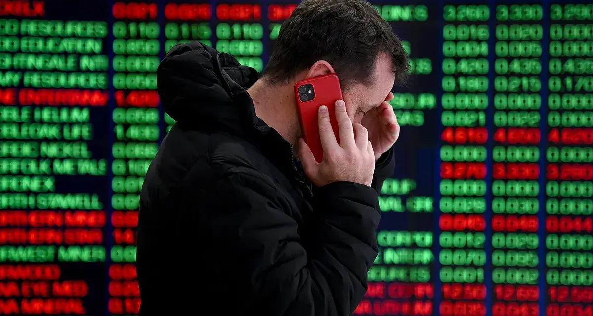Hombre preocupado habla por teléfono frente a pantalla de bolsa de valores de Nueva York, con números rojos y verdes indicando un posible crack.