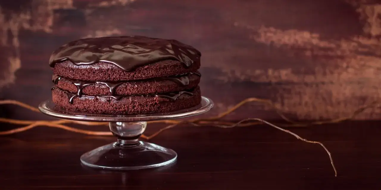 Torta al cioccolato a tre strati, lucida e invitante, su un'alzata di vetro. Gocce di glassa scivolano sui bordi.