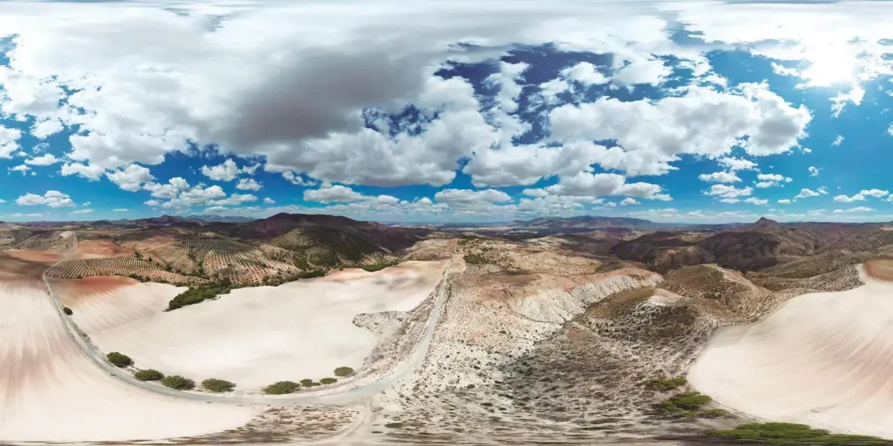 Paisaje árido con colinas, campos cultivados y un camino serpenteante. El cielo azul con nubes blancas contrasta con el desierto de larva.