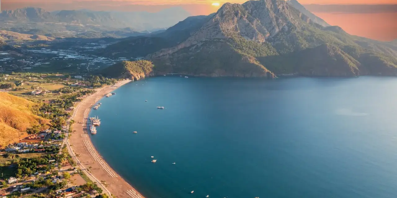 Malownicze wybrzeże Turcji o zachodzie słońca. Góry schodzą do turkusowego morza, a plaża z leżakami i łodziami zaprasza do odpoczynku.