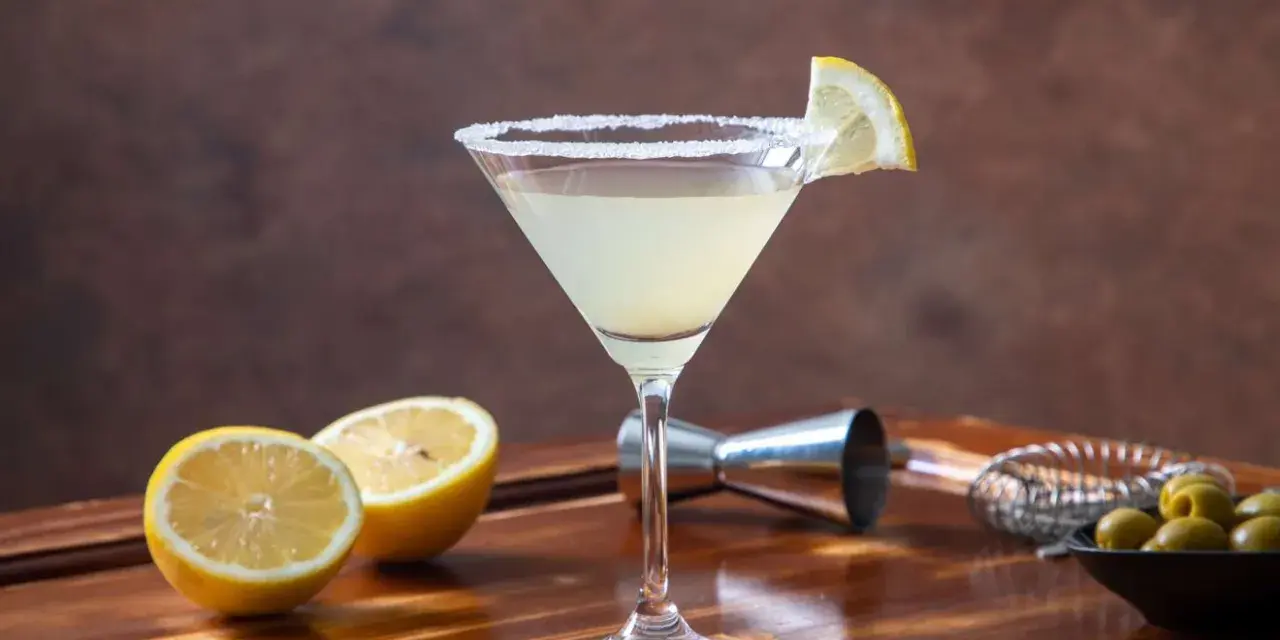 Orzeźwiający drink z martini, udekorowany plasterkiem cytryny i obsypany cukrem. Obok leżą połówki cytryny, oliwki i akcesoria barmańskie. Idealny na domowe drinki z martini.