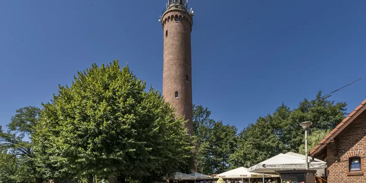 Wysoka, ceglana latarnia morska Sarbinowo góruje nad letnim kurortem z drzewami i ogródkami kawiarnianymi.