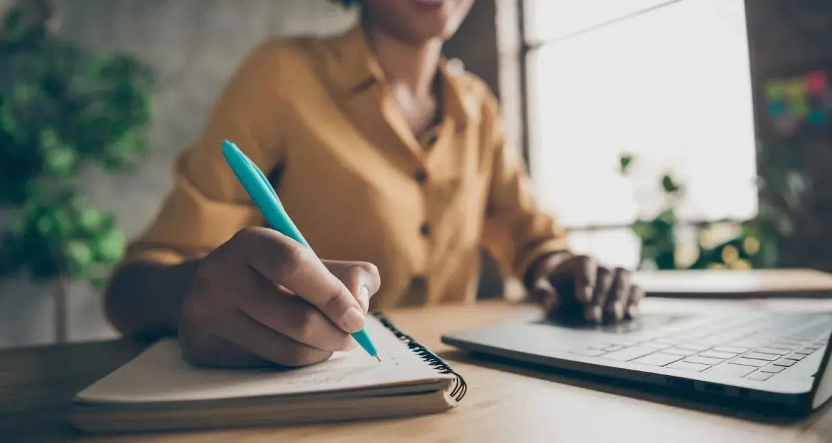 Frau schreibt mit einem blauen Stift Notizen in ein Notizbuch, während sie auf ihrem Laptop tippt. Sie erstellt ein Protokoll ihrer Gedanken.