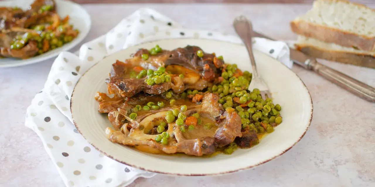 Un piatto di ossobuco con piselli e carote, servito su un piatto bianco con una forchetta. Sullo sfondo, fette di pane.
