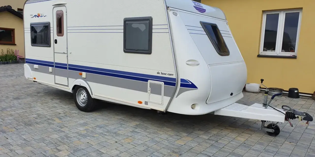 Biała przyczepa kempingowa Hobby 460 UFE "de luxe easy" stoi na kostce brukowej przed żółtym budynkiem.