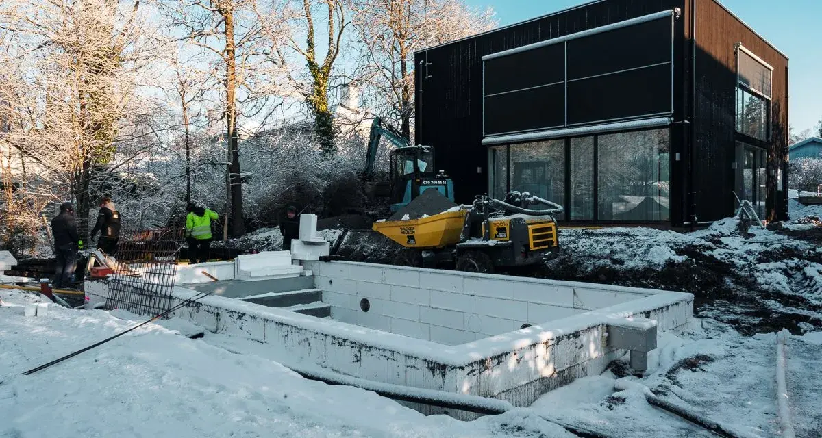 Snö täcker byggarbetsplatsen där en utbyggnad av husets långsida pågår. En grävmaskin och en dumper syns vid en nybyggd poolgrund.