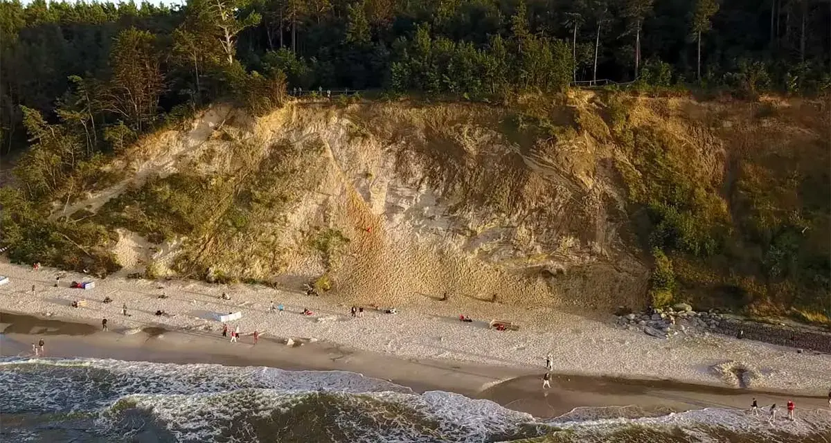 Plaża u stóp najwyższego klifu w Polsce. Ludzie spacerują po piasku, fale obmywają brzeg.