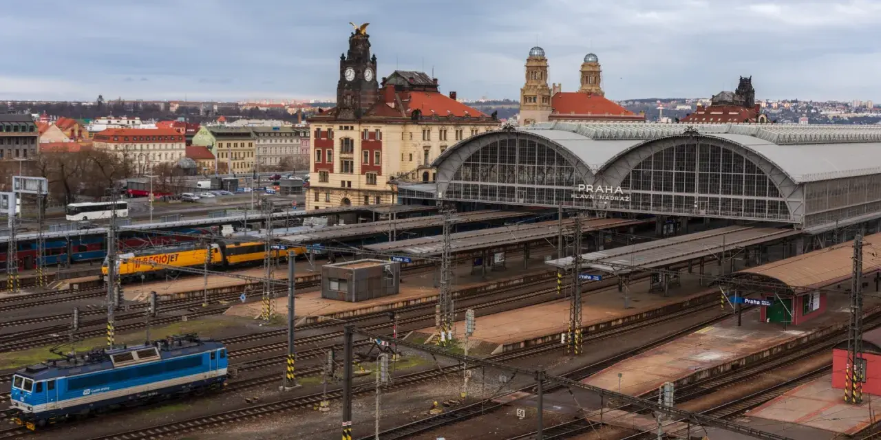 Widok na dworzec kolejowy w Pradze z pociągami na torach i budynkami w tle.