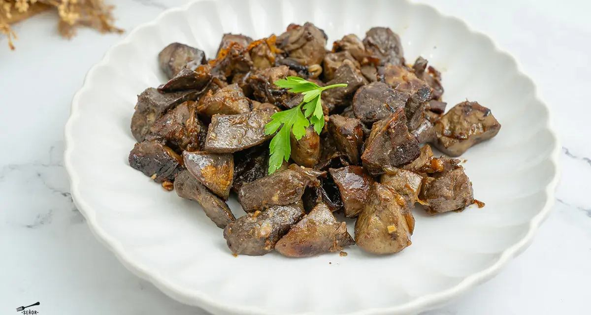 Plato de asadura de cordero salteada, con trozos oscuros y jugosos, adornado con una ramita de perejil fresco.