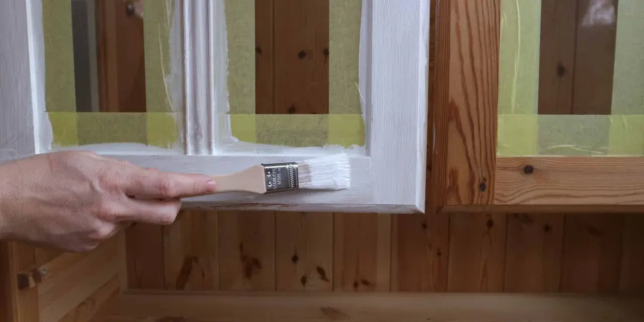 En hand målar en träyta vit med en pensel. Maskeringstejp skyddar intilliggande ytor av furu.