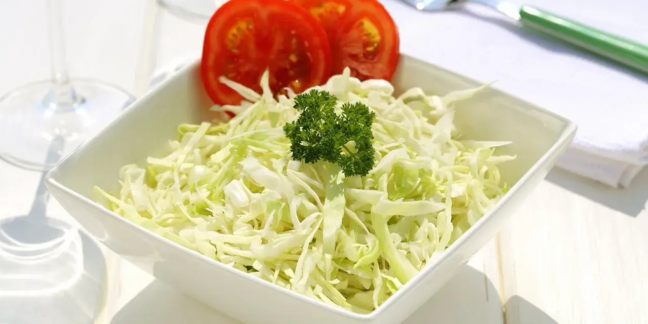 Frischer, selbstgemachter Krautsalat in einer weißen Schüssel, garniert mit Petersilie und Tomatenscheiben. Daneben liegen ein grüner Löffel und eine Serviette.