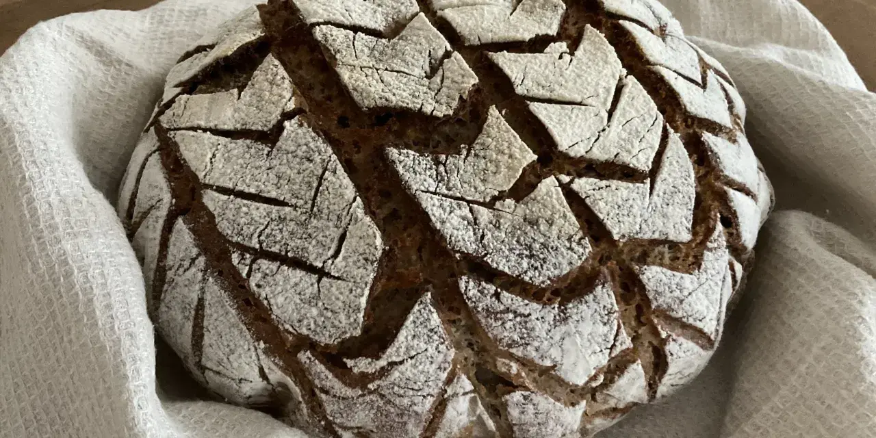 Ein rustikales, rundes buchweizenbrot sauerteig mit einer gemusterten Kruste, bestäubt mit Mehl, liegt auf einem weißen Leinentuch.