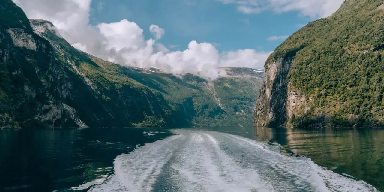 Rejs po Geirangerfjordzie. Pogoda sprzyja, chmury wiszą nad zielonymi zboczami gór, a za statkiem widać biały ślad na spokojnej wodzie.