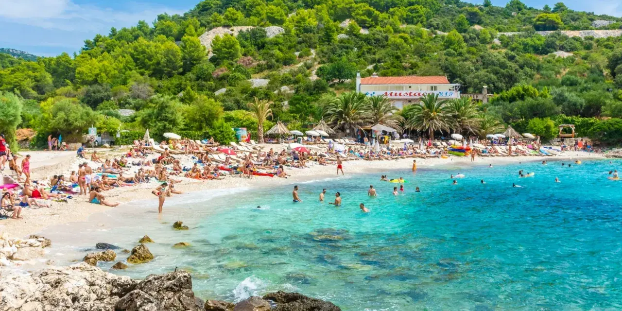 Słoneczna plaża z turkusową wodą, mnóstwem ludzi odpoczywających na leżakach i kąpiących się w morzu. W tle zielone wzgórza i budynek z czerwoną dachem. Idealne miejsce na pierwszy minutę wakacji.
