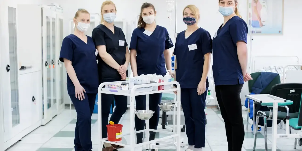 Pięć studentek pielęgniarstwa w granatowych uniformach i maseczkach stoi przy wózku medycznym. To ich przyszła praca dla studenta pielęgniarstwa.