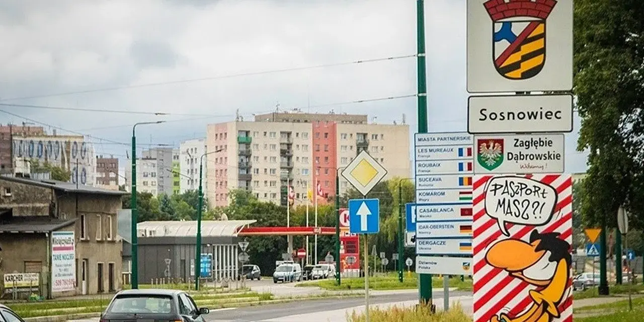 Znak drogowy "Sosnowiec" i humorystyczny napis "Paszport masz?!" przy wjeździe do miasta. W tle bloki mieszkalne.